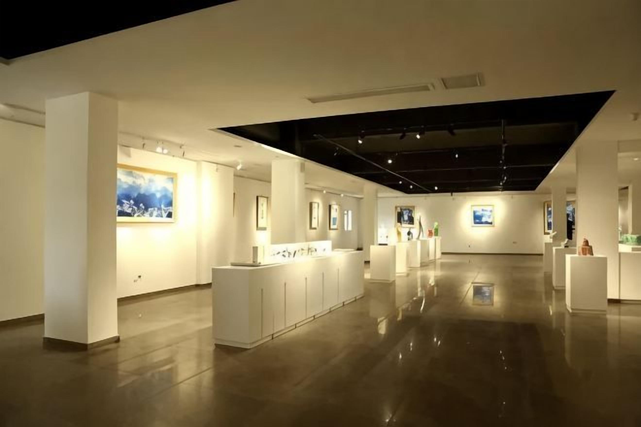 Una amplia galería con paredes blancas y suelo pulido, que presenta varias exhibiciones de arte y un elemento de iluminación central.