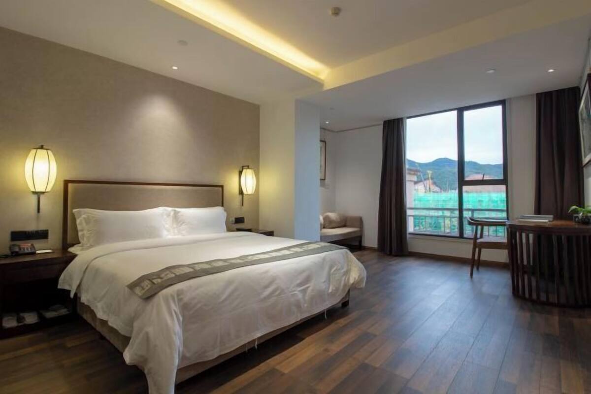 A hotel room with a large bed, bedside lamps, a desk with a chair, and a view of the outdoors through a large window.