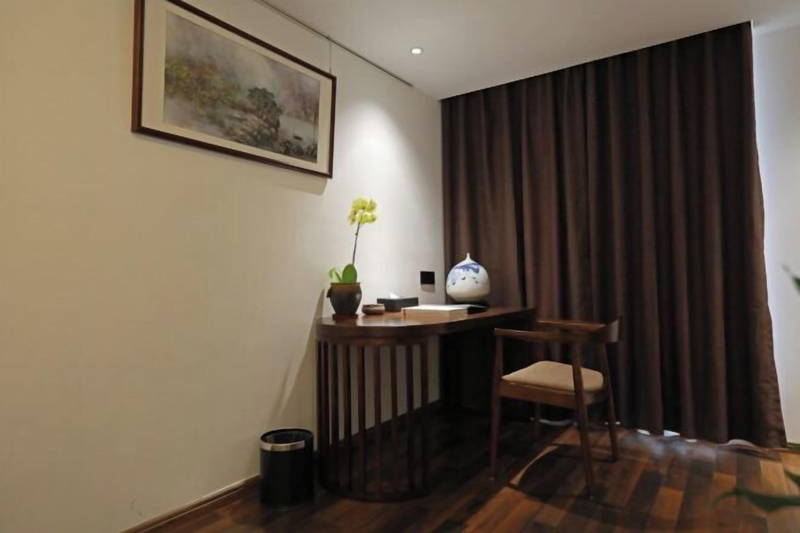 A hotel room with a wooden floor, a desk with a vase and a potted plant, a chair, a painting on the wall, and a window with curtains.