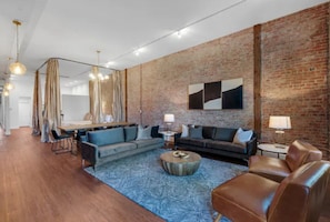 A modern living room with a brick wall, a blue patterned rug, leather sofas, and a dining area with a table and chairs.