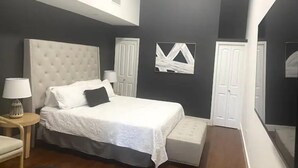 Ein Schlafzimmer mit einem großen Bett, einem Nachttisch mit Lampe, einem Sessel und einem kleinen Hocker. Es gibt zwei Türen und ein an der Wand befestigtes Kunstwerk.