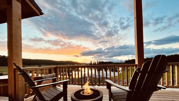 A wooden deck with two Adirondack chairs and a fire pit, overlooking a lake and forested landscape during sunset. - Clydesdale Outpost (Whitefish)