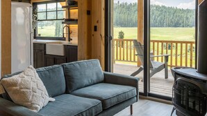A cozy cabin interior with a wooden ceiling, a sofa with a cushion, a dining table, and a view of a forest through large windows. - Clydesdale Outpost (Whitefish)