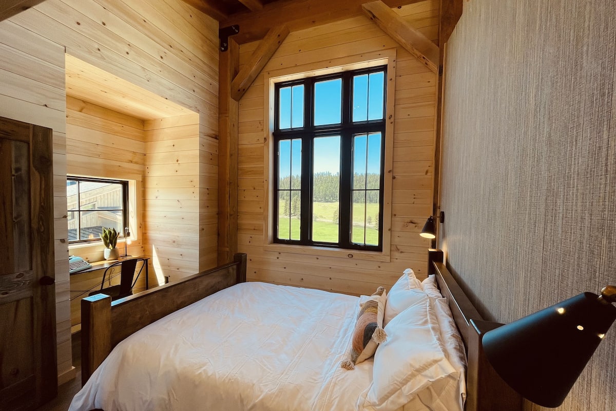 A wooden cabin bedroom with a large window, a bed, a desk, and a chair.