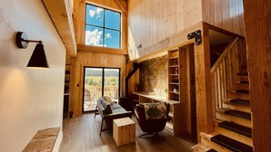 A spacious wooden interior with a high ceiling, a large window offering a scenic view, a cozy seating area, and a staircase. - Clydesdale Outpost (Whitefish)
