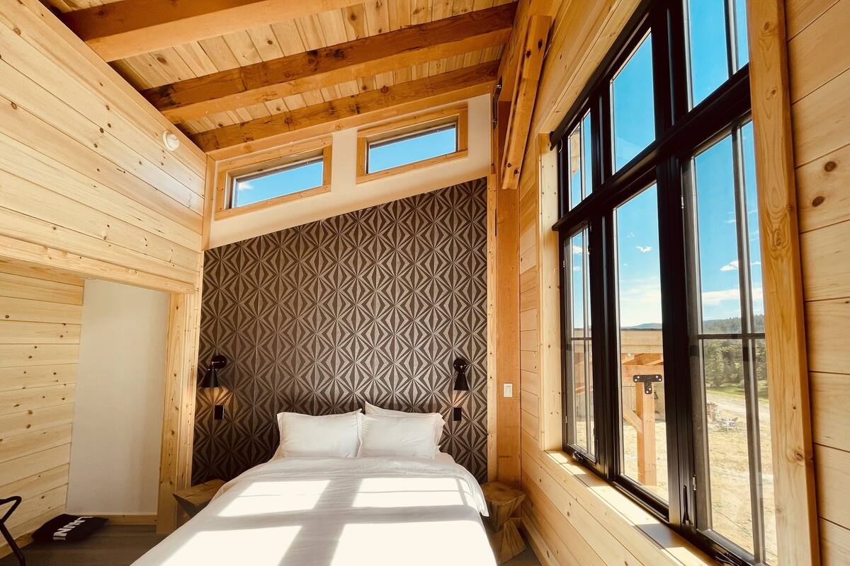 A wooden cabin bedroom with a bed, two skylights, a patterned wall, and a large window.