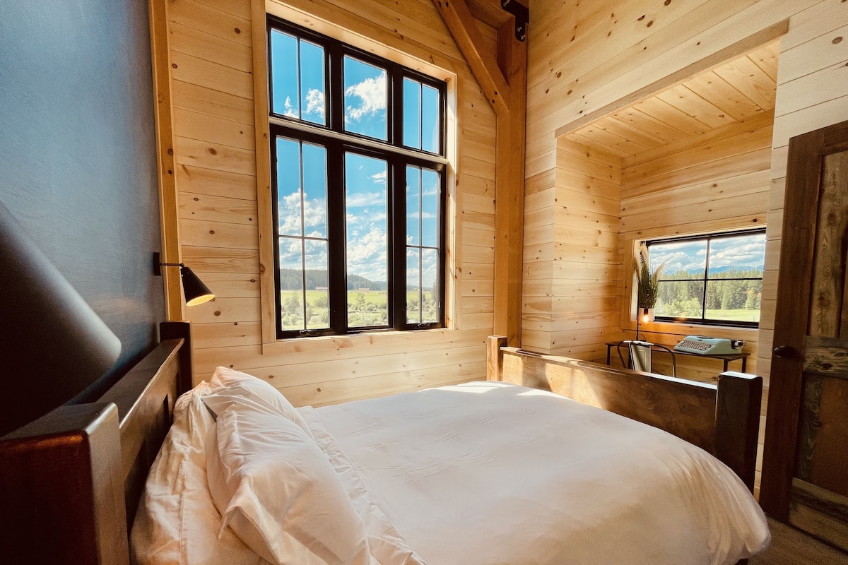 A wooden cabin bedroom with a large bed, a desk with a typewriter, and a view of the outdoors through a large window.