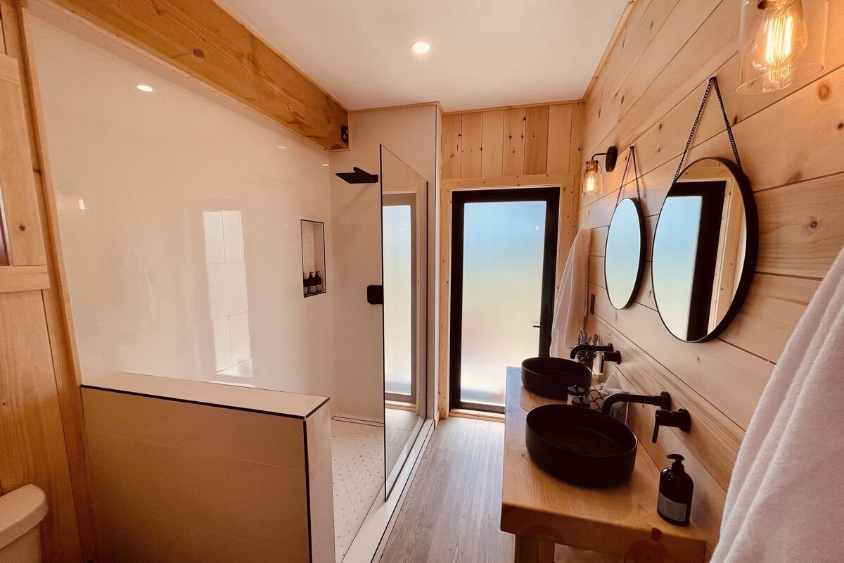 A modern bathroom with wooden walls, a glass shower enclosure, a sink with a black basin, and two round mirrors.