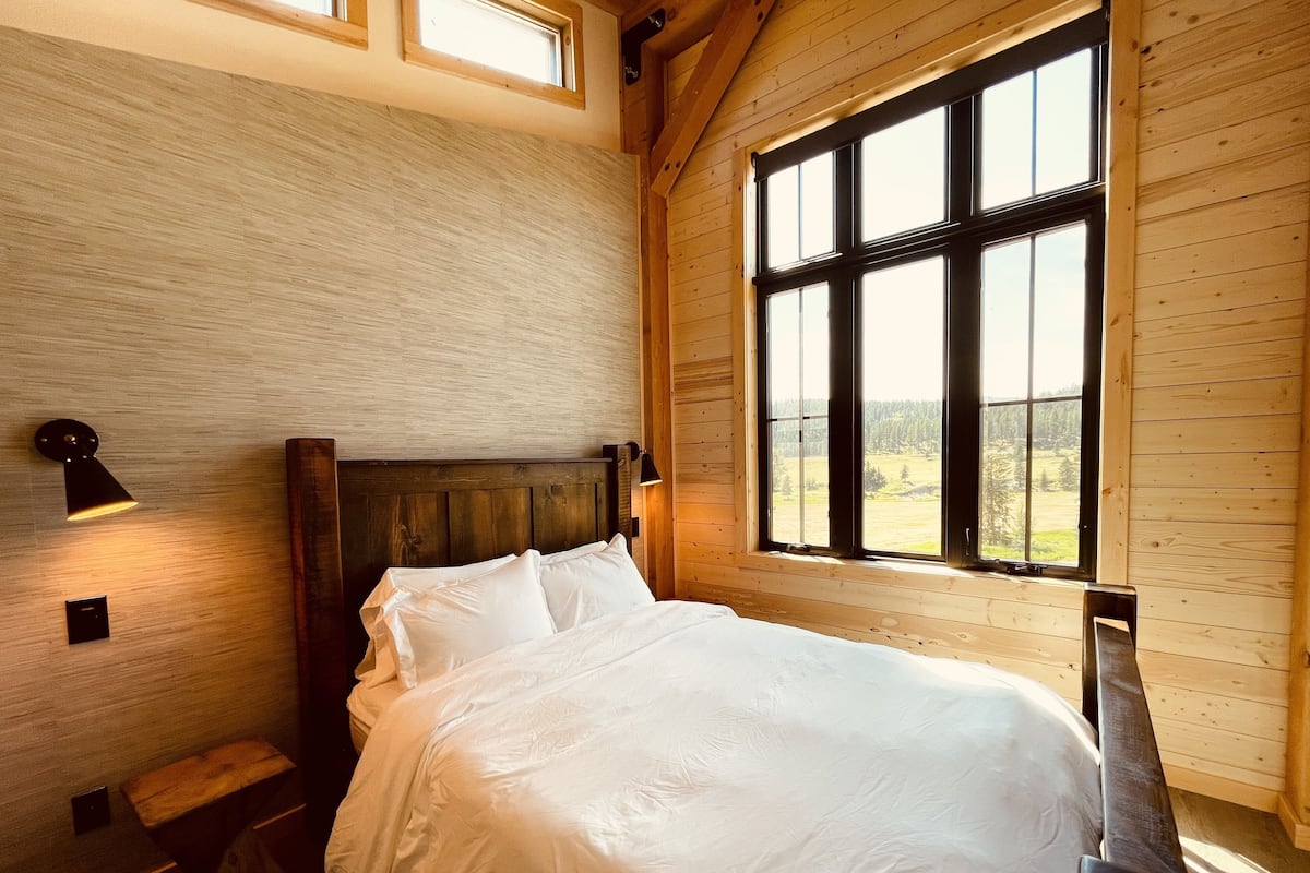 A bedroom with a wooden headboard, a large window with a view of a forest, and two wall-mounted lamps.