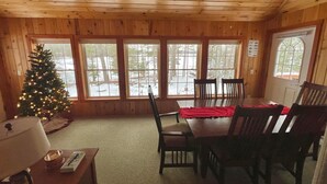 Interior acogedor de una cabaña con techo de madera, una mesa de comedor con sillas, un árbol de Navidad y ventanas grandes que ofrecen vistas a un paisaje nevado.