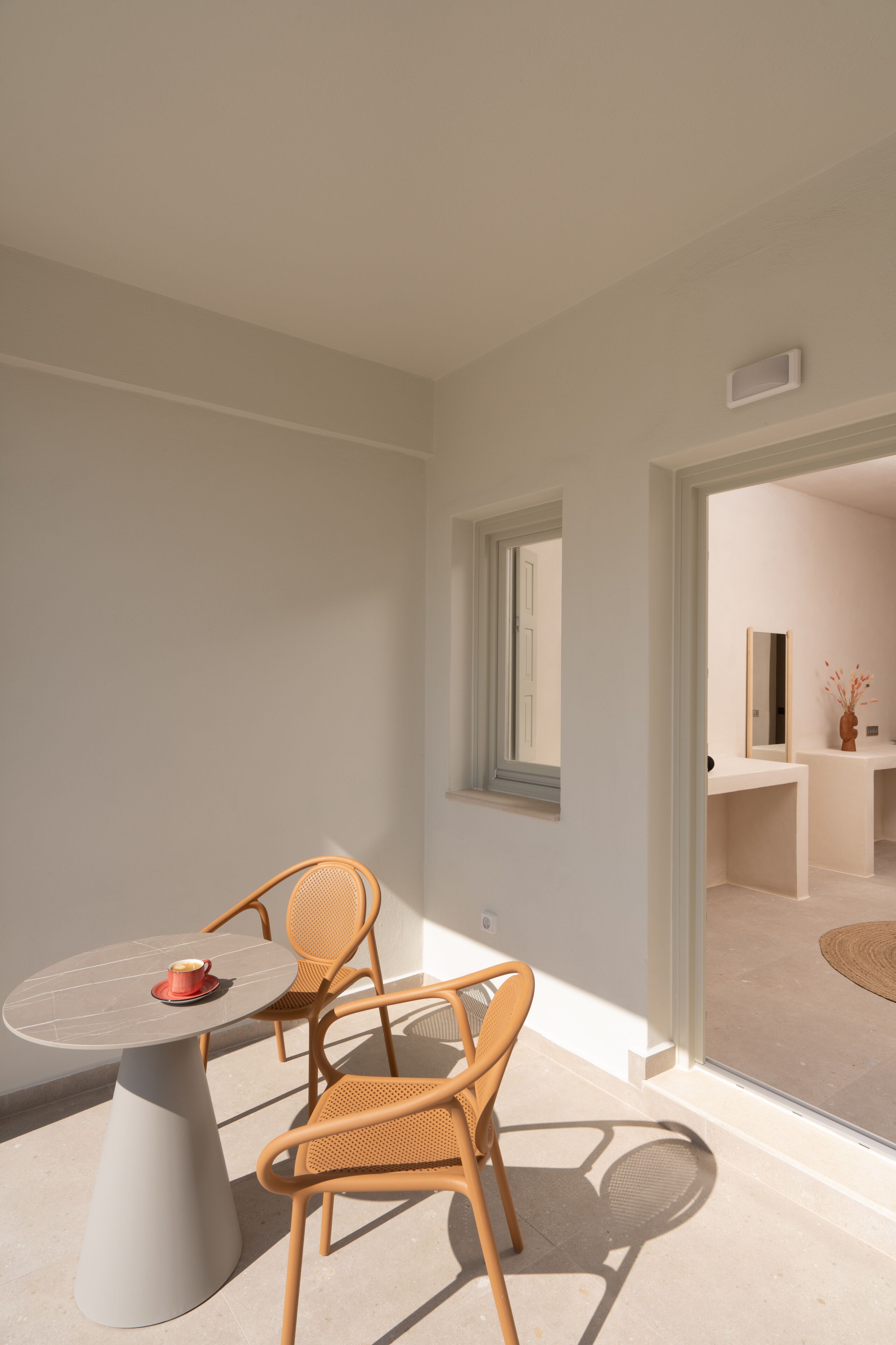 A modern room with a round table and two wicker chairs, a small window, and a view into another room.
