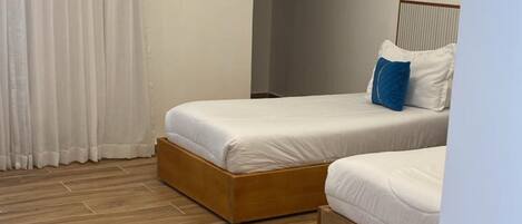 A modern bedroom with two single beds, wooden flooring, a large window with white curtains, and a ceiling light.