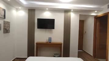 A hotel room with a large bed, a flat-screen TV mounted on the wall, a wooden bedside table with a bottle and glass, and a trash bin.