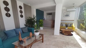 A modern living room with a teal sofa, a glass coffee table, and a yellow sofa. There are potted plants and a staircase leading to an upper level.