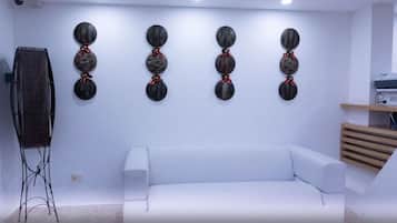 Ein modernes Wohnzimmer mit einem weißen Sofa, einem Couchtisch und einem an der Wand montierten Fernseher. An den Wänden sind dekorative Kunstwerke angebracht und ein Holzregal ist mit Gegenständen bestückt.