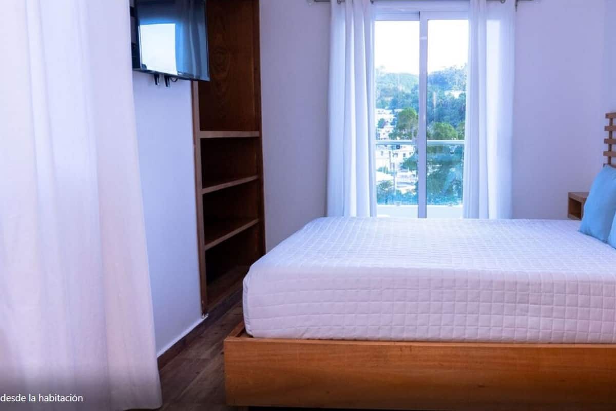 Un dormitorio con cama de madera, ropa de cama blanca y vista a un paisaje urbano a través de un ventanal grande.