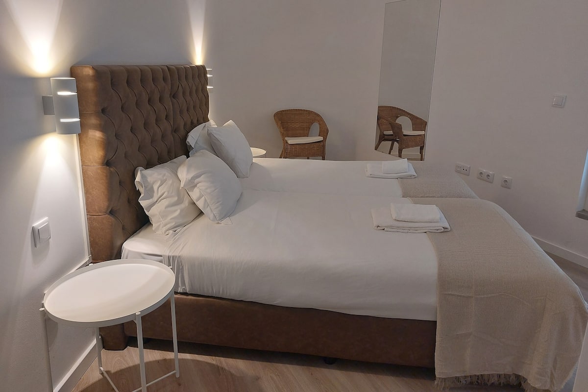 A neatly made bed with white linens and pillows, a round white side table, and a wooden headboard.