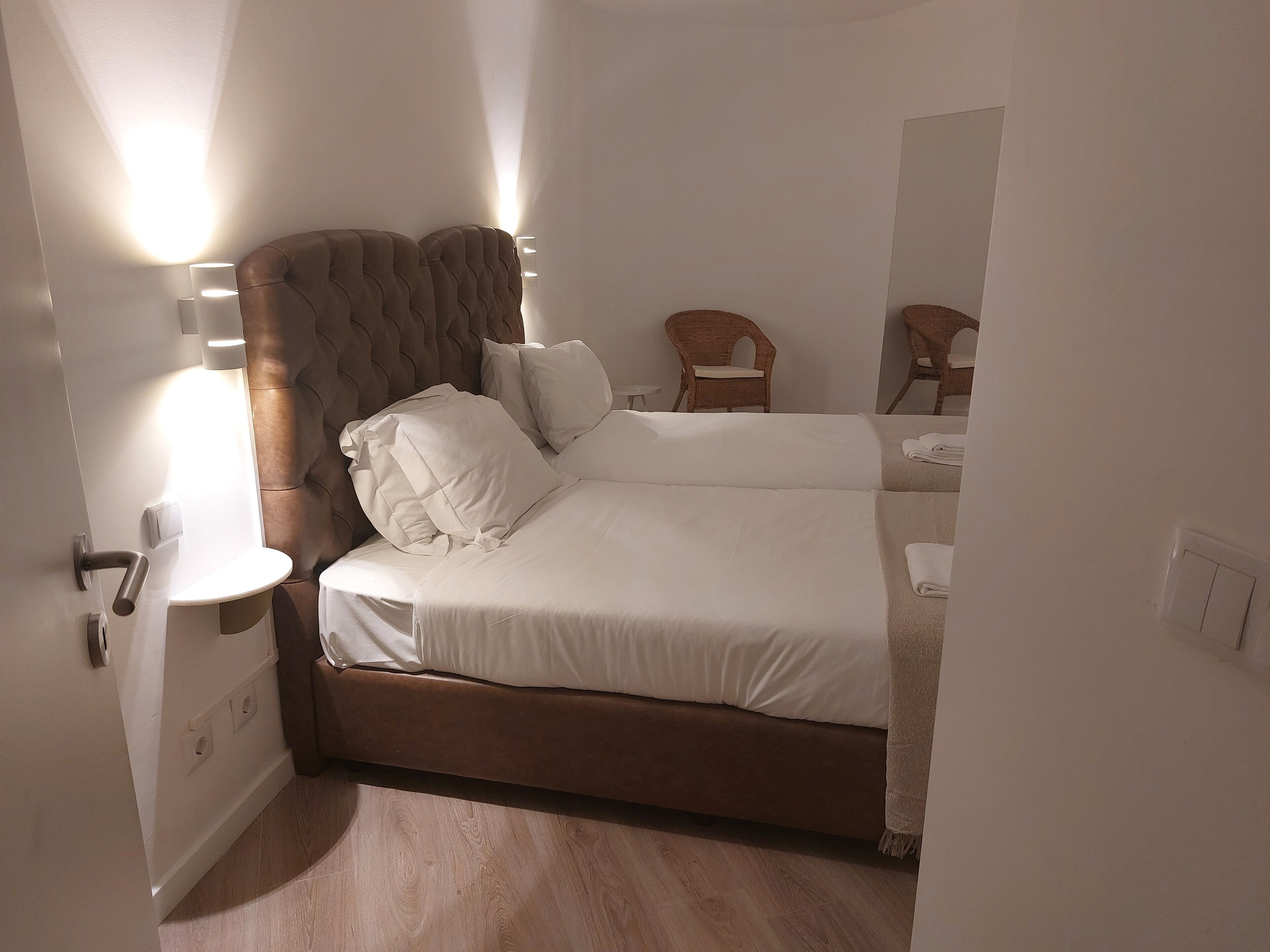 Een slaapkamer met een tweepersoonsbed, nachtkastje, stoel en een wandlamp.
