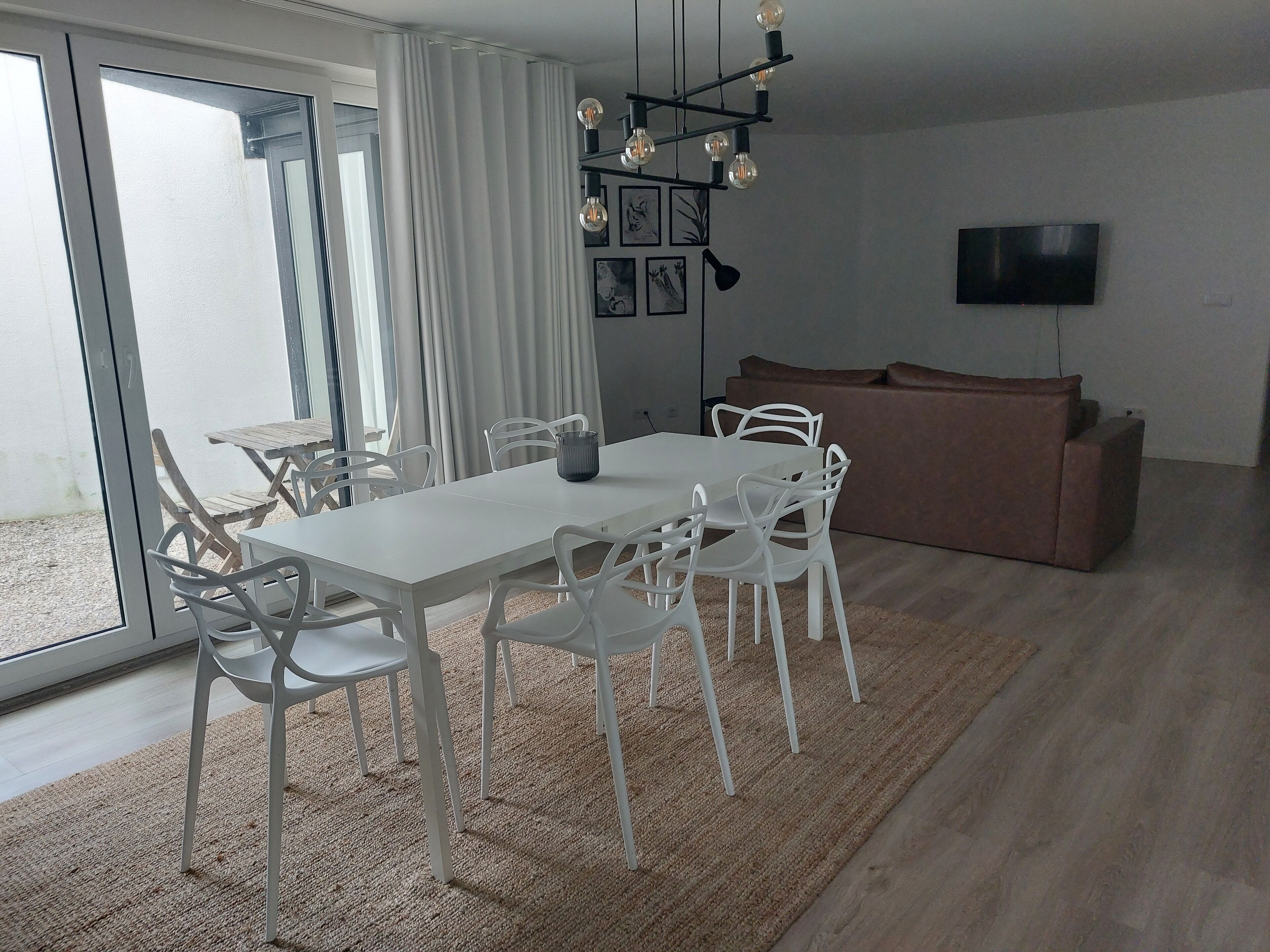 A modern living room with a dining area, featuring a white table and chairs, a brown sofa, a flat-screen TV, and a chandelier.