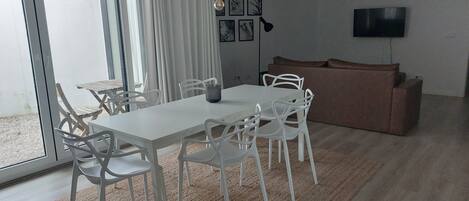 A modern living room with a dining area, featuring a white table and chairs, a brown sofa, a flat-screen TV, and a chandelier.