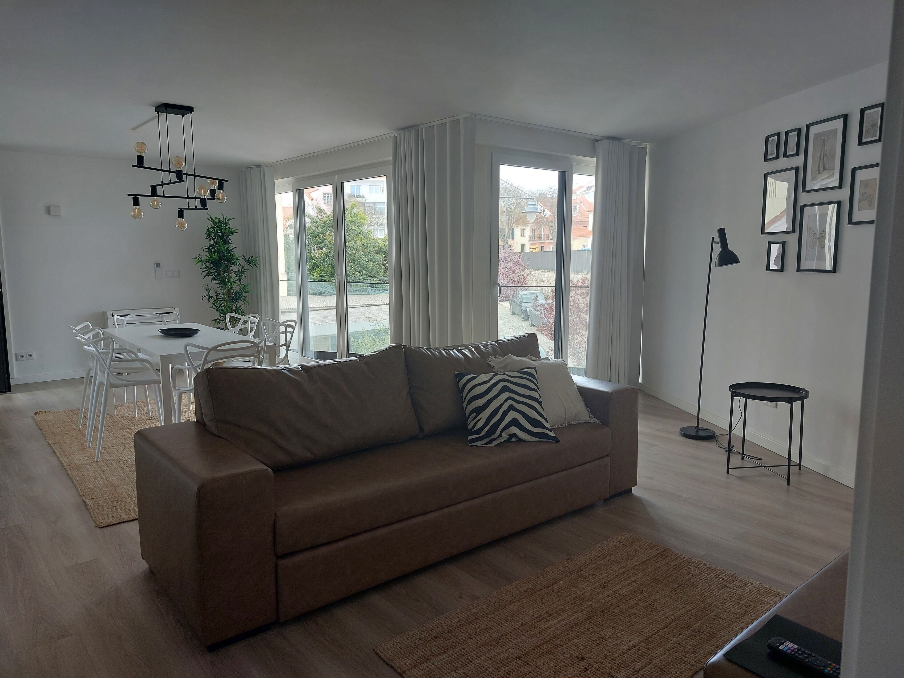 Una sala moderna con un sofá marrón, una mesa de comedor blanca y sillas. Hay un ventanal con cortinas, una lámpara de pie y cuadros en la pared.