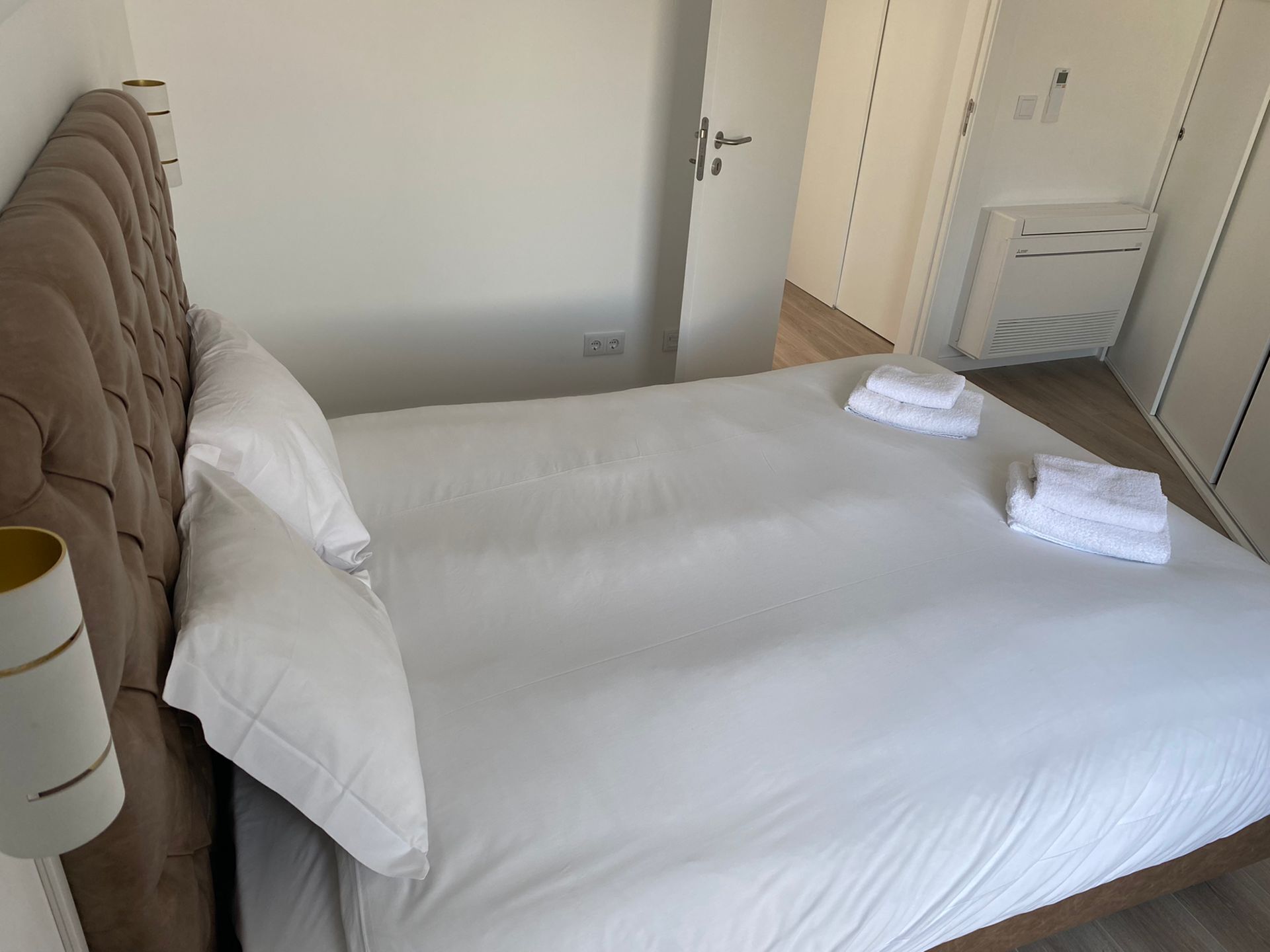 Un lit bien fait, avec du linge de lit blanc et deux serviettes pliées, une tête de lit capitonnée et un mur blanc orné d’une porte et d’une petite armoire blanche.