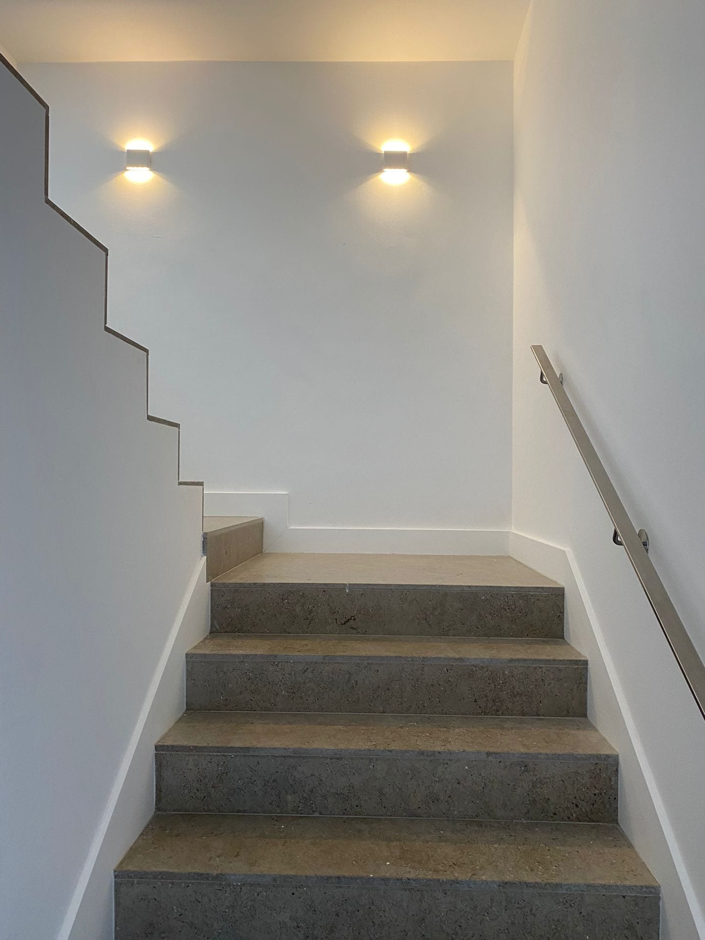 Una escalera de hormigón con barandilla metálica que lleva a un rellano con dos luces empotradas en la pared.