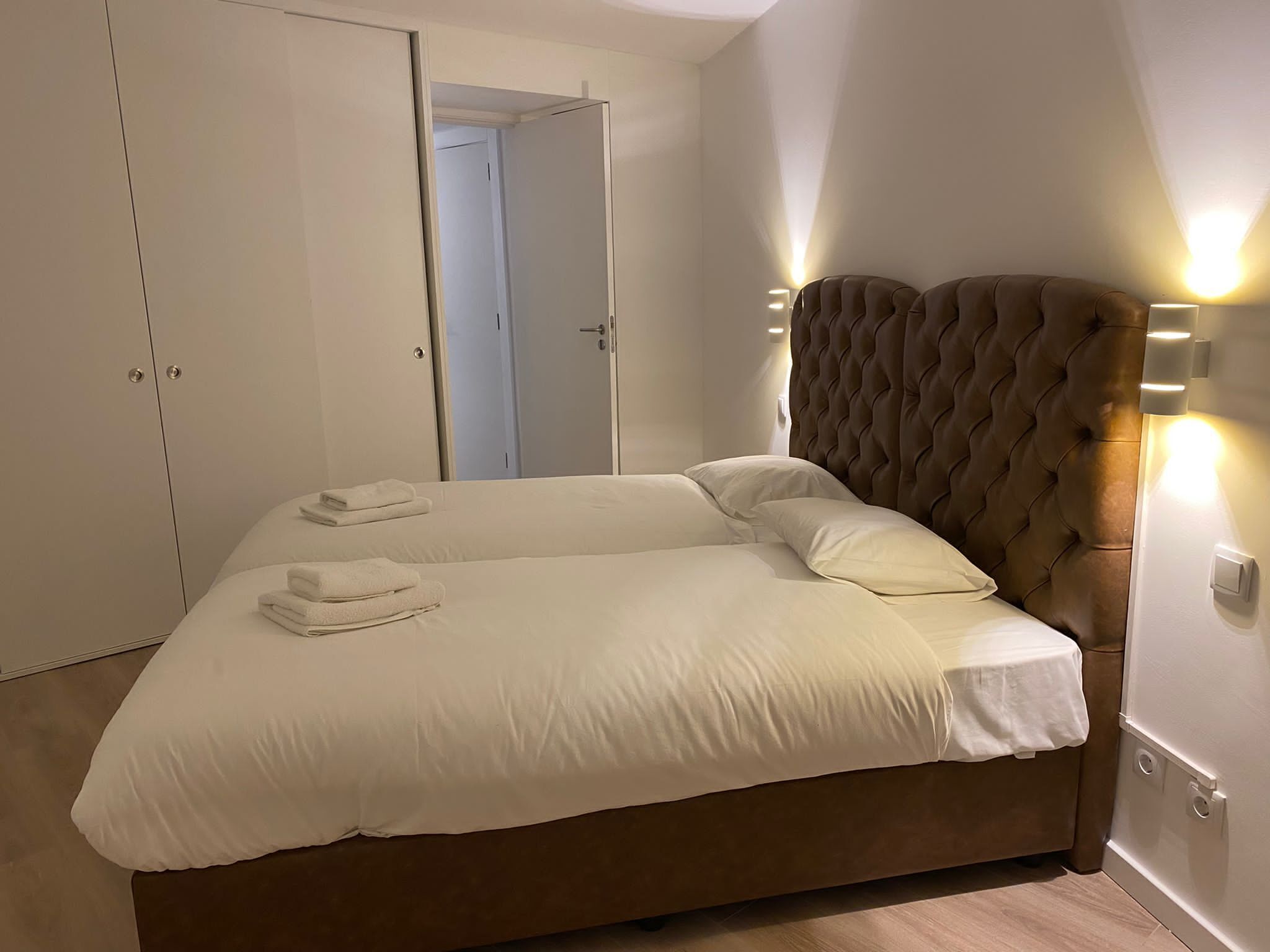 A neatly made bed with white linens and two folded towels, a tufted headboard, and a wall-mounted light fixture.