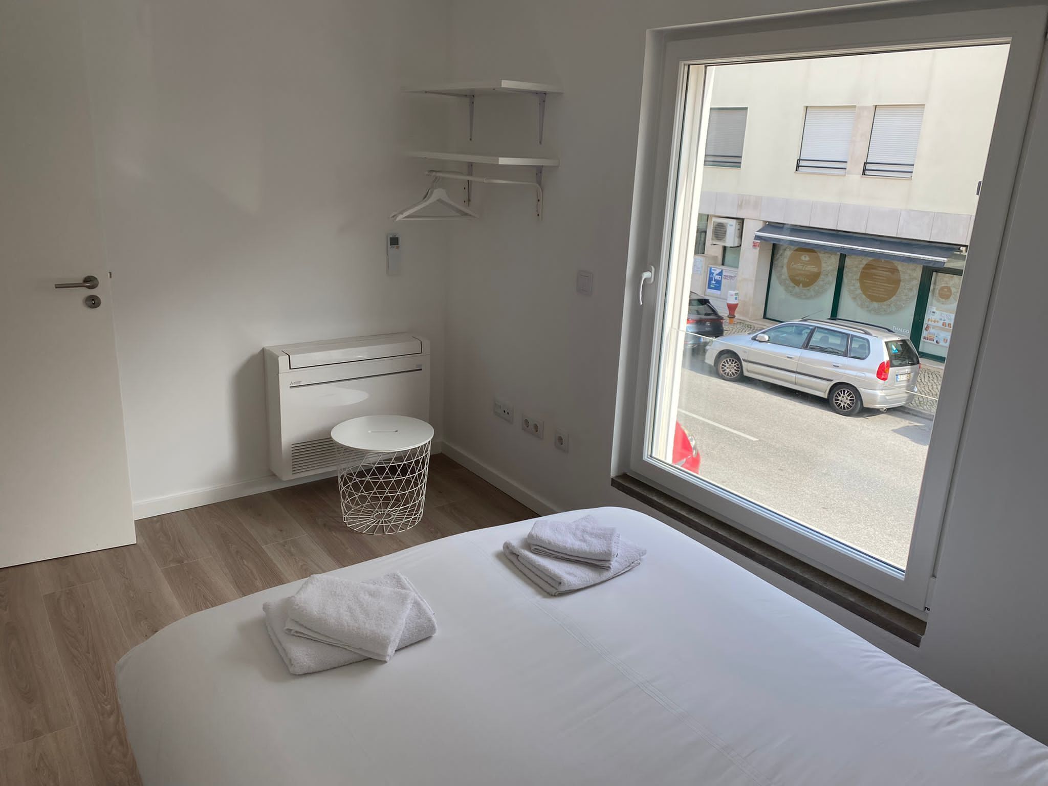 A hotel room with a bed, a round white trash bin, a white heater, and a window with a view of a street and parked cars.