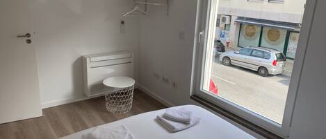 A hotel room with a bed, a round white trash bin, a white heater, and a window with a view of a street and parked cars.