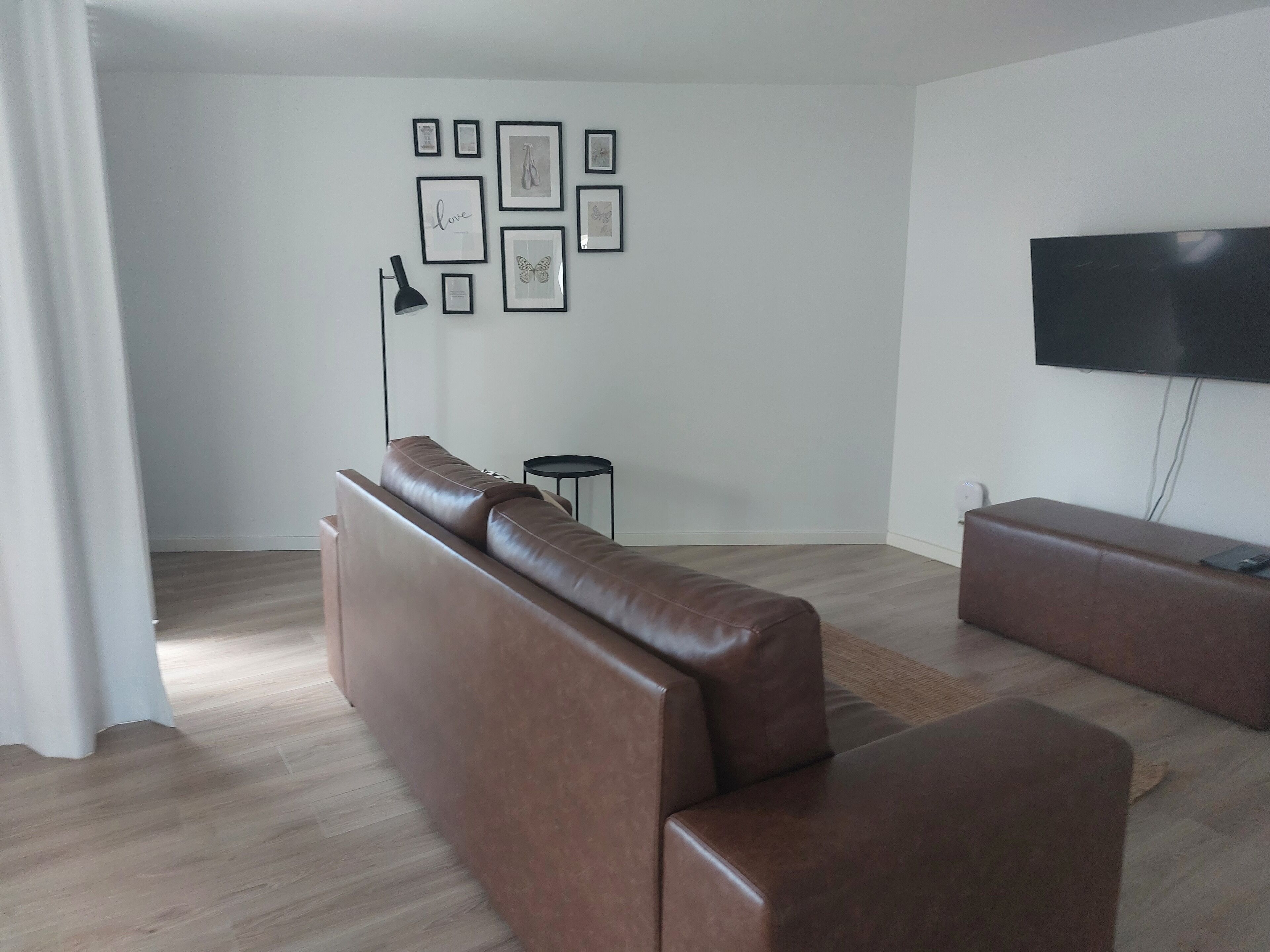 A modern living room with a brown leather sofa, a small round table, a floor lamp, and a flat-screen TV mounted on the wall.