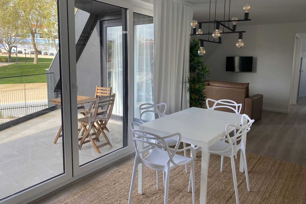 A modern dining area with a white table and chairs, a large sliding glass door leading to a balcony, and a black chandelier.