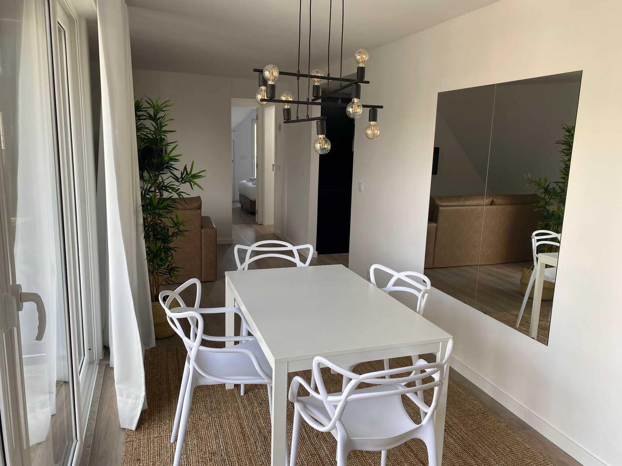 Een eetgedeelte met een witte tafel en stoelen, een moderne kroonluchter en een grote spiegel aan de muur.