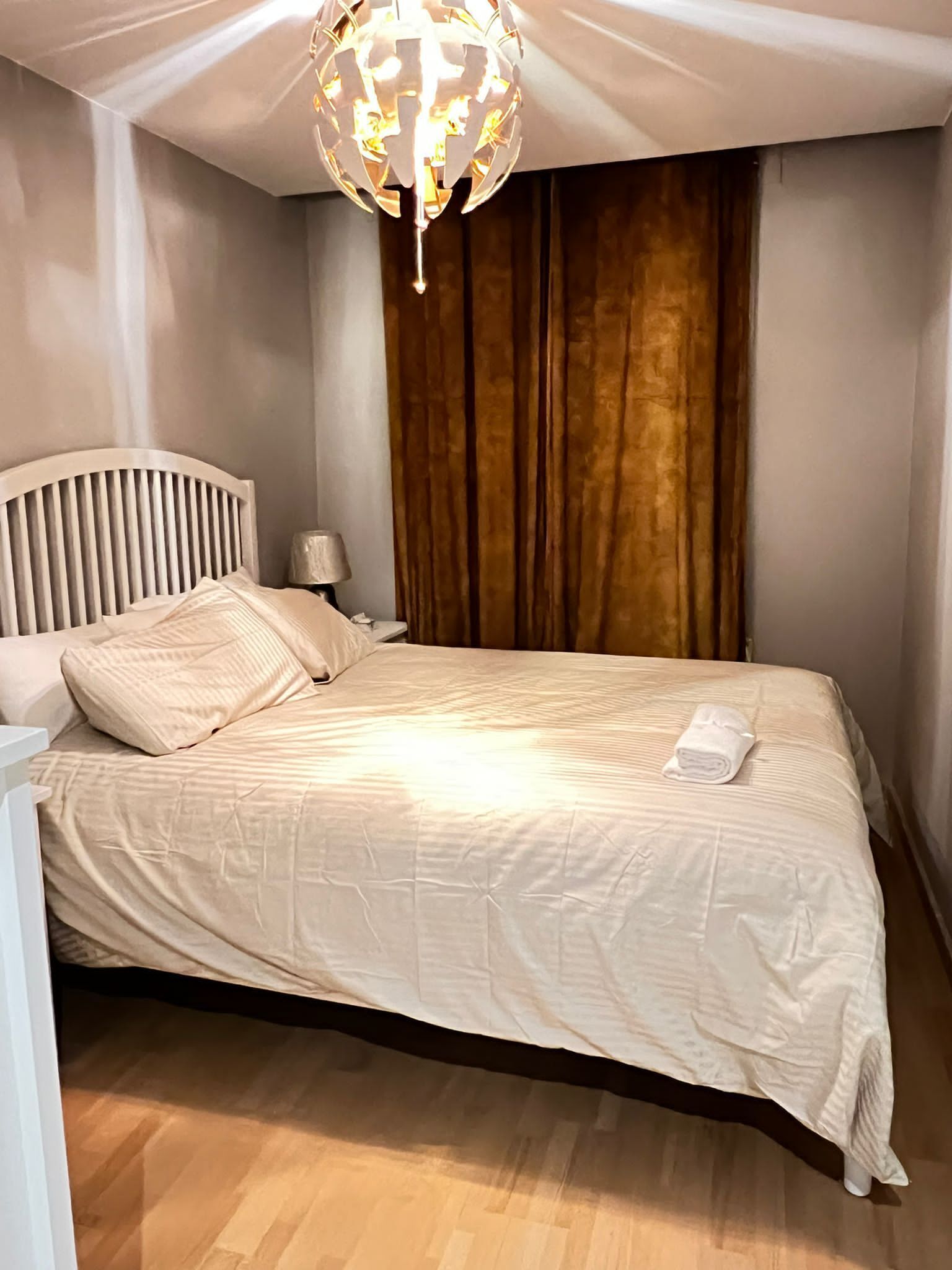 A neatly made bed with a white comforter and pillows, a bedside lamp, a wooden headboard, and a hanging light fixture.