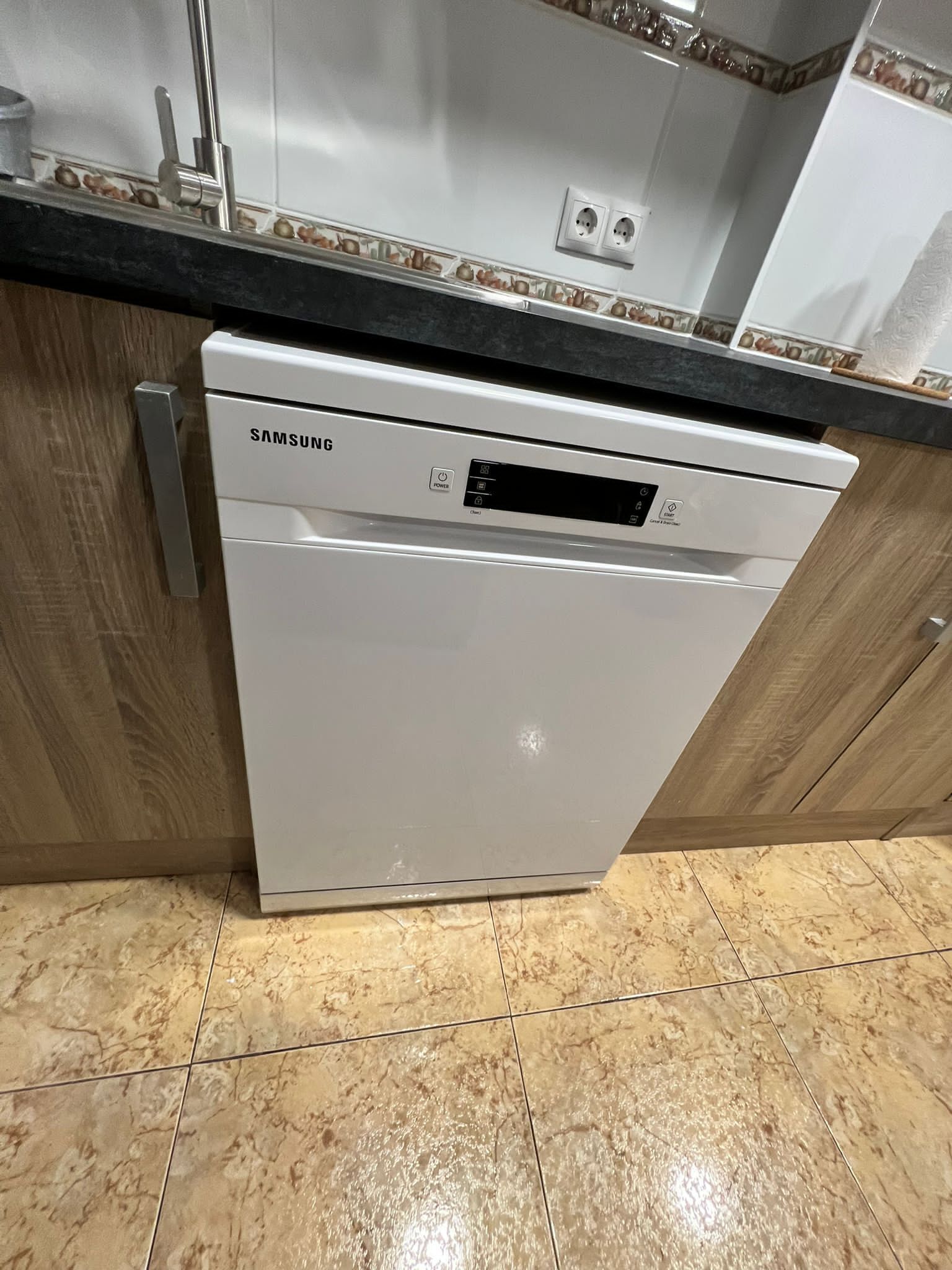 A Samsung dishwasher in a kitchen with wooden cabinets and a marble floor.
