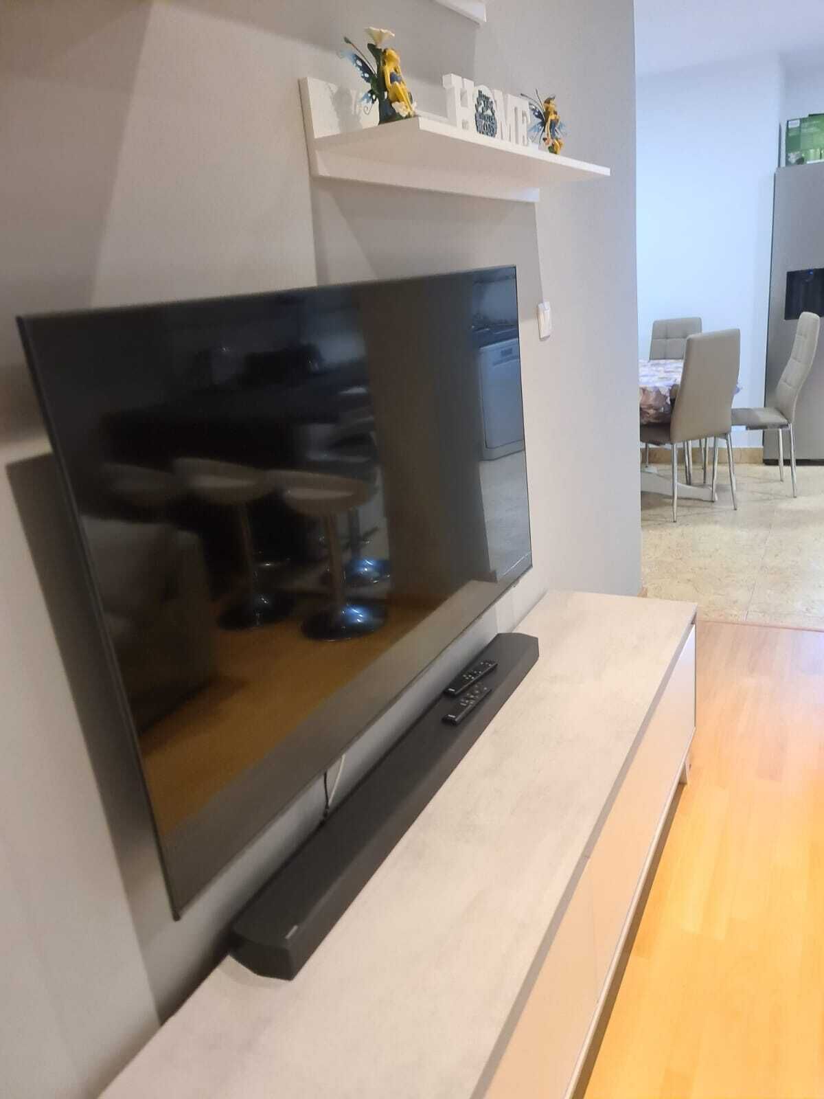 Una sala moderna con un televisor de pantalla plana montado en la pared, una mesa de centro de madera y una zona de comedor con sillas y una mesa al fondo.