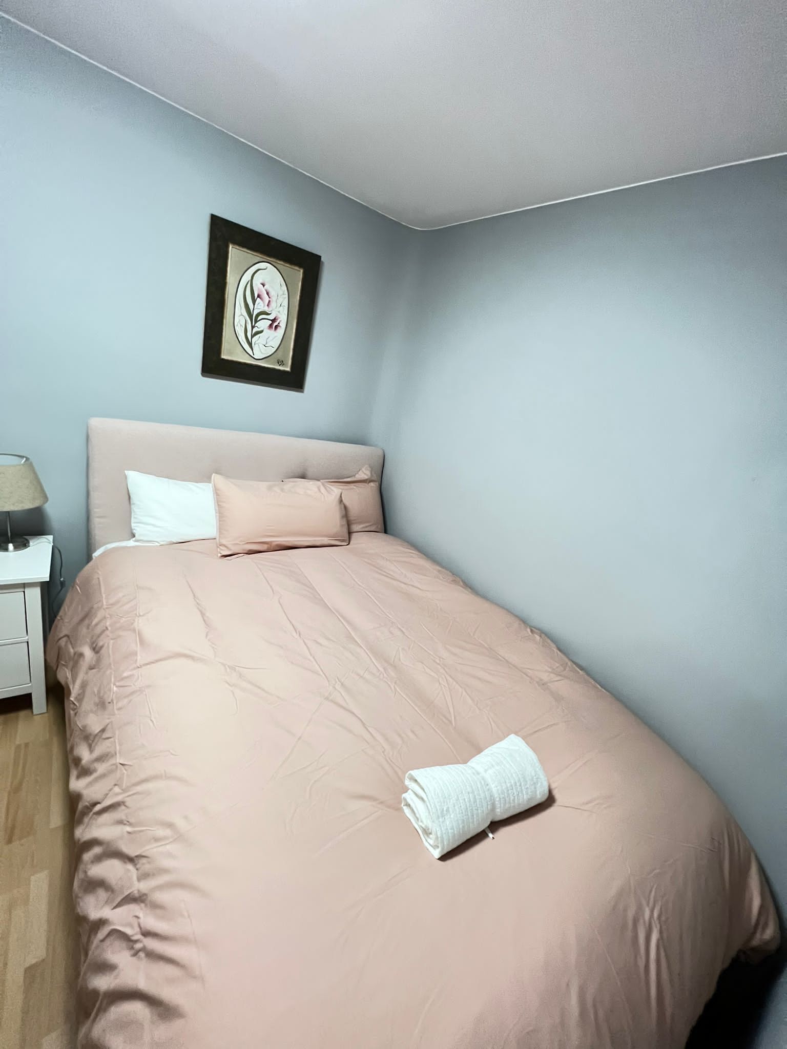Una cama bien hecha con colcha y almohadas rosadas, una obra de arte enmarcada en la pared y una mesita de noche blanca con una lámpara.