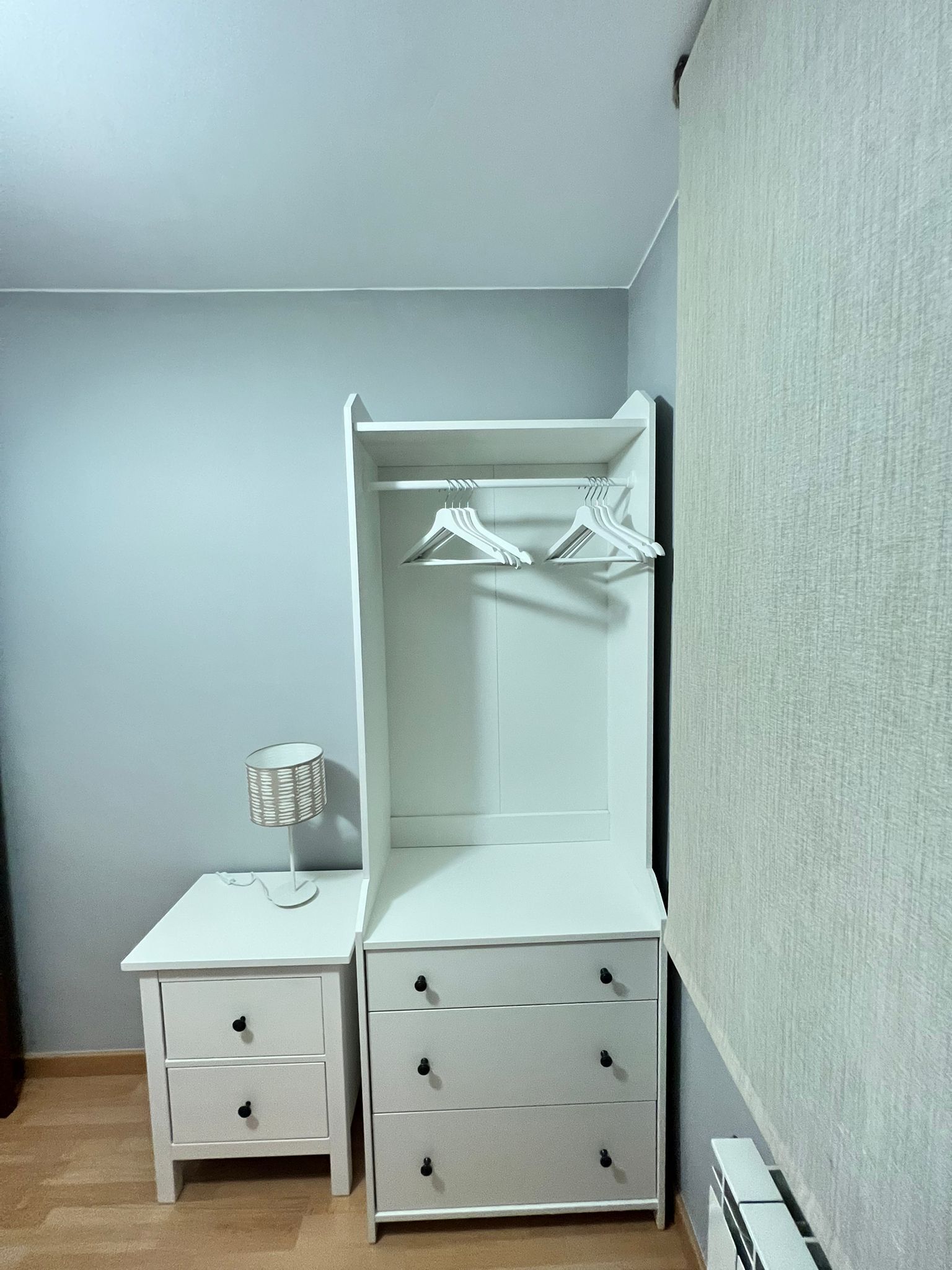 Een klein, wit nachtkastje met een lamp en een dressoir met drie lades en een spiegel.