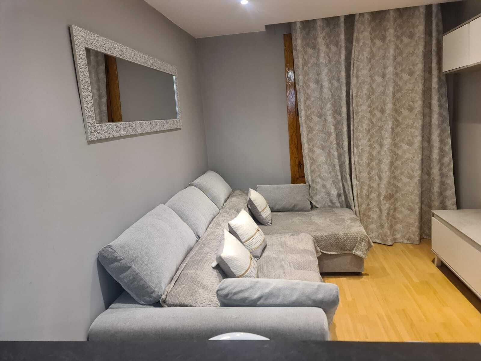 Un salon moderne avec un canapé d’angle gris, un plancher en bois, un meuble blanc et un rideau à motifs.
