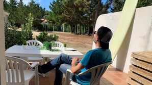 Un homme est assis à une table blanche, entouré de chaises, sur une terrasse en plein air dont le sol est recouvert de carreaux. L’endroit est agrémenté de verdure et bordé d’un mur blanc.