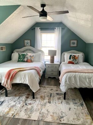 Een slaapkamer met twee bedden, een plafondventilator, een raam met gordijnen en een klein nachtkastje met een lamp.