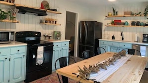 Una cocina con armarios blancos, estufa negra, refrigerador y una mesa de comedor con un arreglo decorativo de hojas.