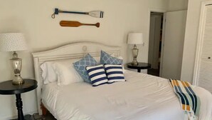 Una cama bien tendida con ropa blanca, dos almohadas a rayas azules y blancas, y una sola almohada azul. Hay dos mesitas de noche con lámparas, una a cada lado de la cama. Un remo de madera está montado en la pared sobre la cama.