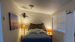 Un dormitorio con una cama, dos lámparas de noche, una lámpara de techo, una ventana con persianas y una obra de arte colgada en la pared.