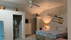 Een slaapkamer met een bed, een stoel, een nachtkastje, een lamp, een plafondventilator en een klein plankje met decoratieve spullen.