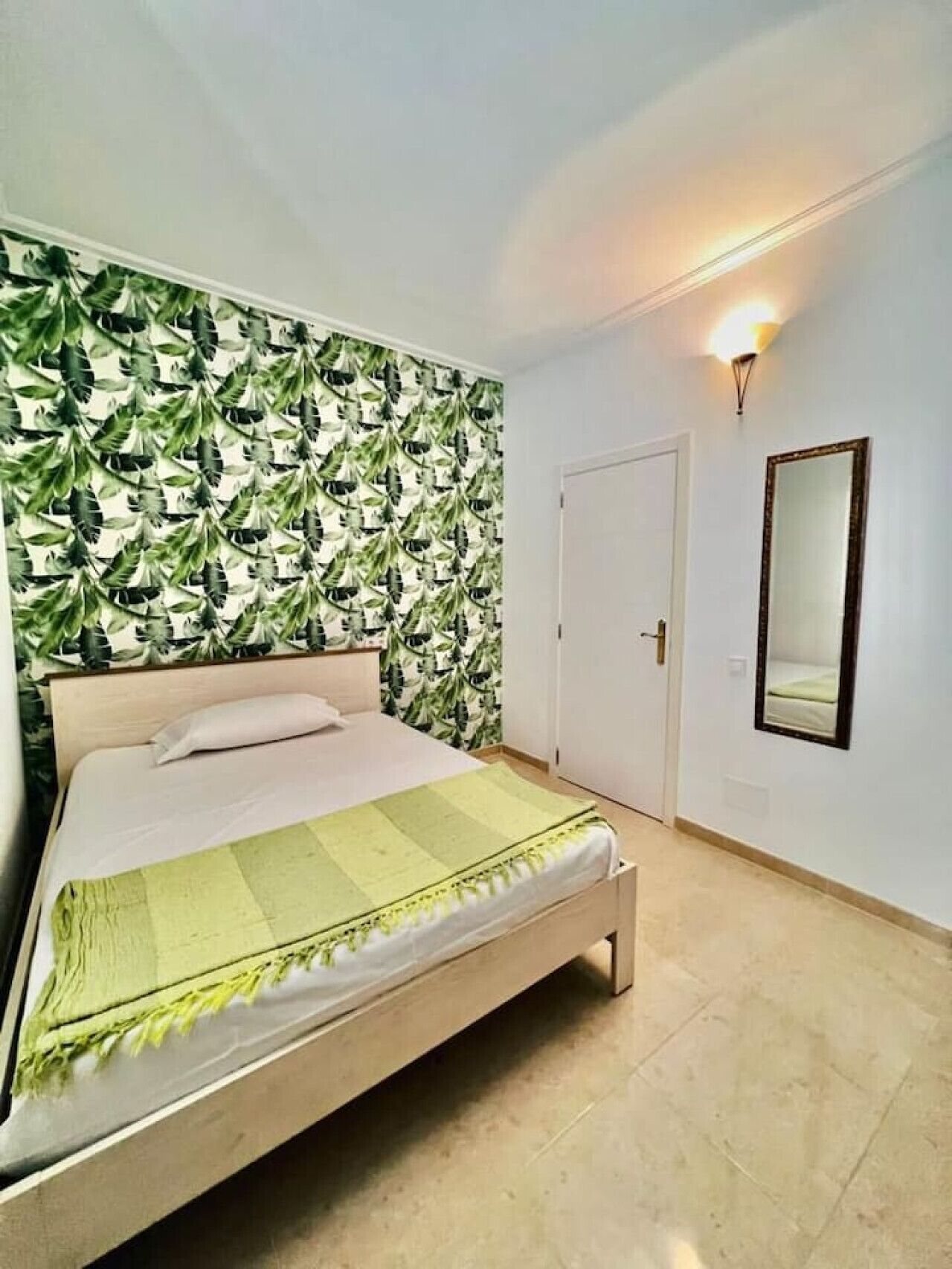 A bedroom with a bed, a wall with a leafy pattern, a door, and a wall-mounted mirror.