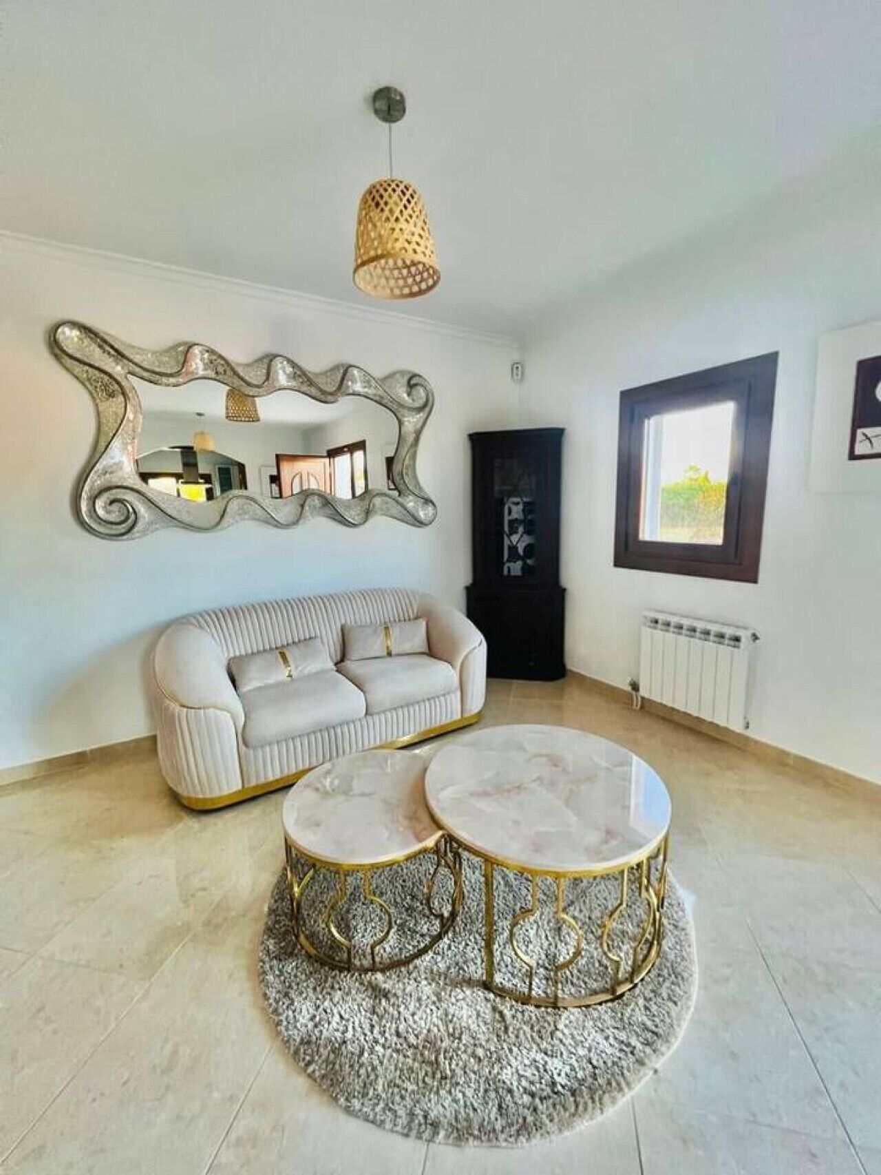 Una sala de estar moderna con un sofá, dos mesas redondas de centro, un espejo decorativo, una lámpara colgante y un ventanal con vistas.