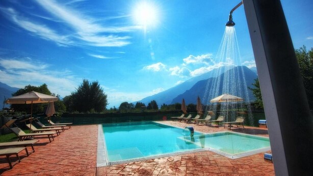 A pool area with lounge chairs and umbrellas, a water feature, and mountains in the background. - Agriturismo Alla Poncia - Monolocale 3 (Gravedona)