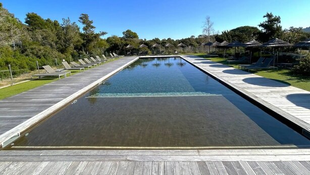 A long swimming pool with a wooden deck, surrounded by lounge chairs and umbrellas.