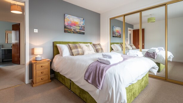 A bedroom with a large bed, a nightstand with a lamp, a painting on the wall, and a mirror with a gold frame. - Centrally located, modern & comfortable apartment with parking. (Inverness)